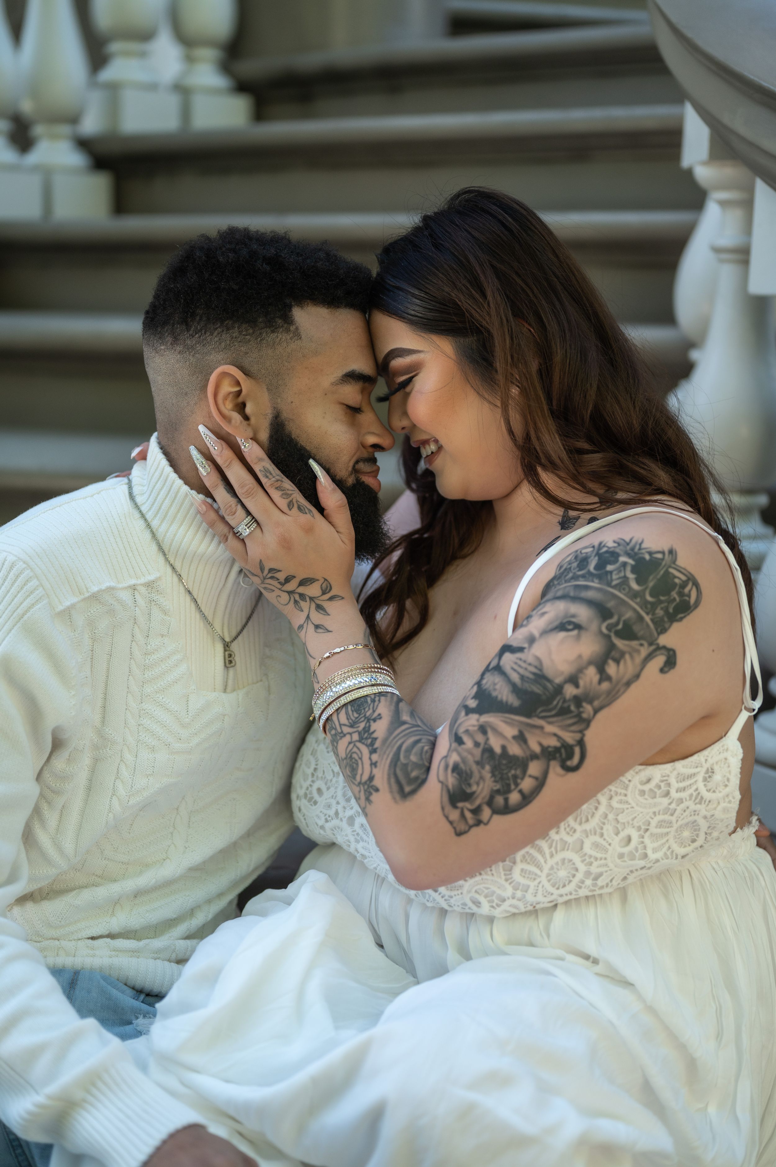 A couple shares a tender moment on stone steps, both wearing white with visible tattoos.