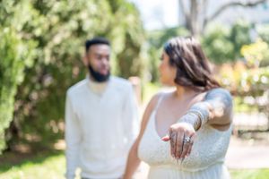 A couple in wedding attire shows off an engagement ring in a sunlit garden setting.