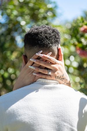 Tattooed hands with long white nails and a wedding ring embrace the back of a head.
