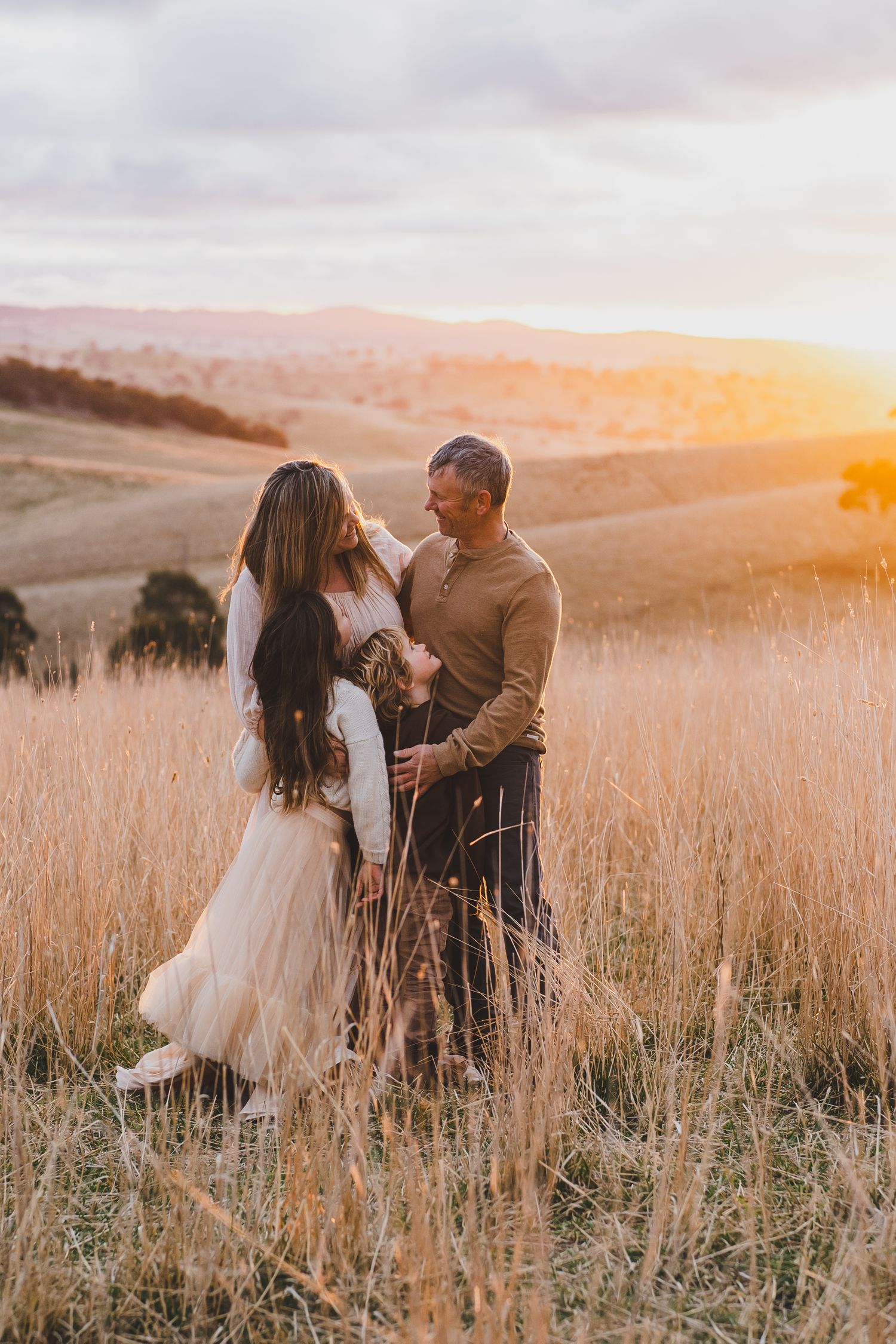 Family of three embracing in golden tall grass field at sunset with rolling hills behind.