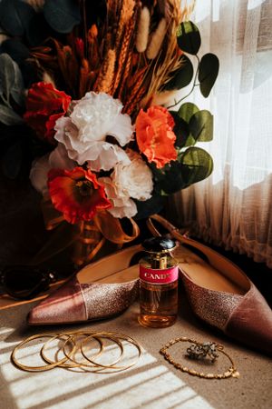 Wedding accessories including glittery shoes, perfume, jewelry and floral arrangement in warm lighting.