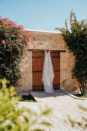 A white wedding dress hangs on stone wall between wooden doors framed by pink bougainvillea flowers.