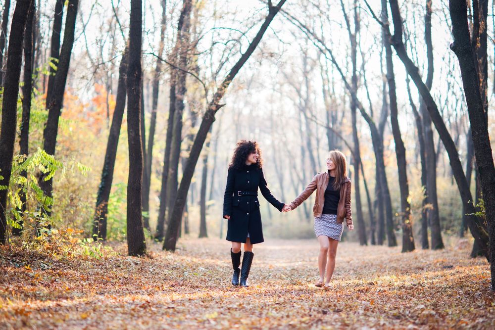 Two friends holding hands while walking through a wooded path covered in fall leaves.