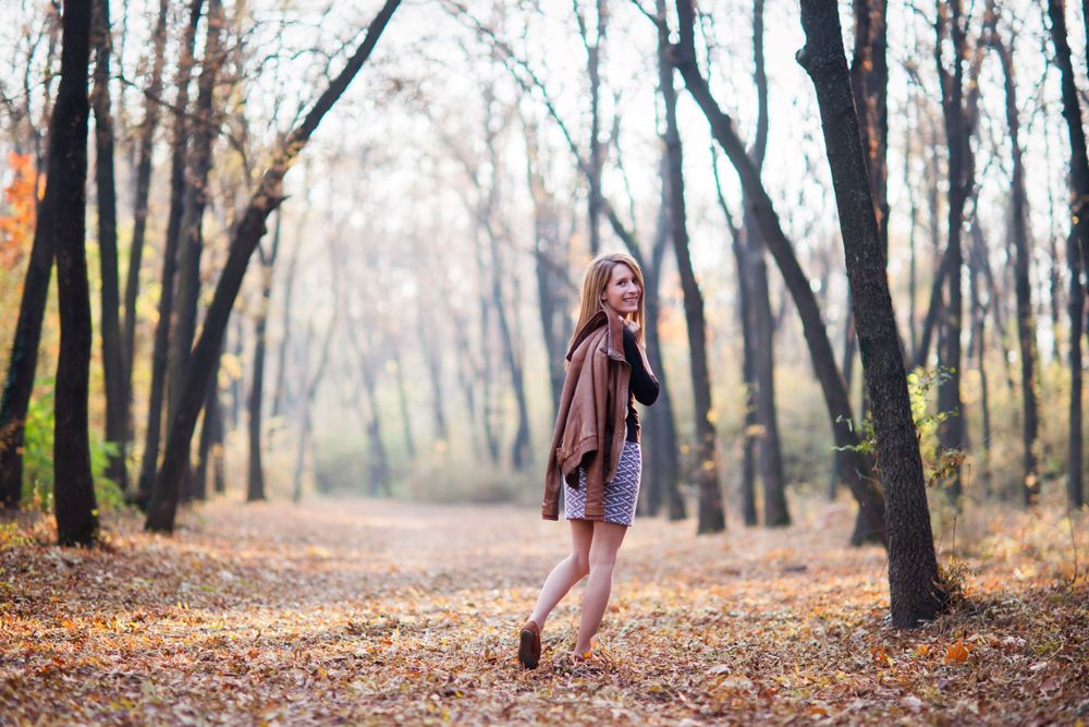 Person in brown coat poses playfully among autumn trees on forest path.