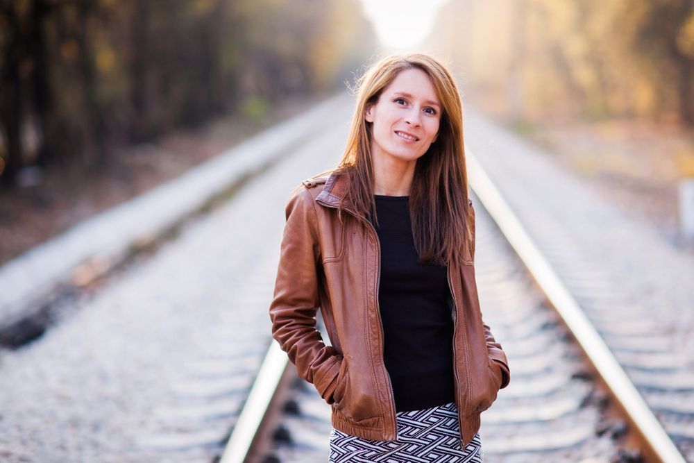 Person in leather jacket and black top posing on railroad tracks with autumn foliage in background.