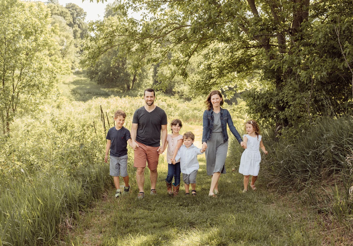 Family of six with four children walking hand in hand along a sunny green nature trail.