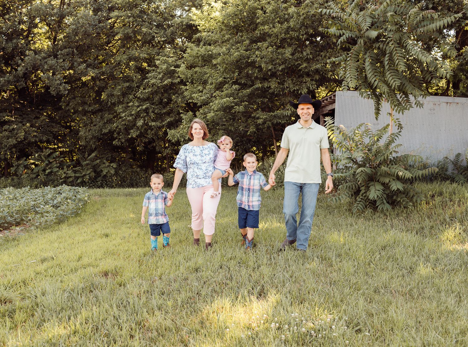 Happy family of five holding hands walking through a lush green backyard on a sunny day.
