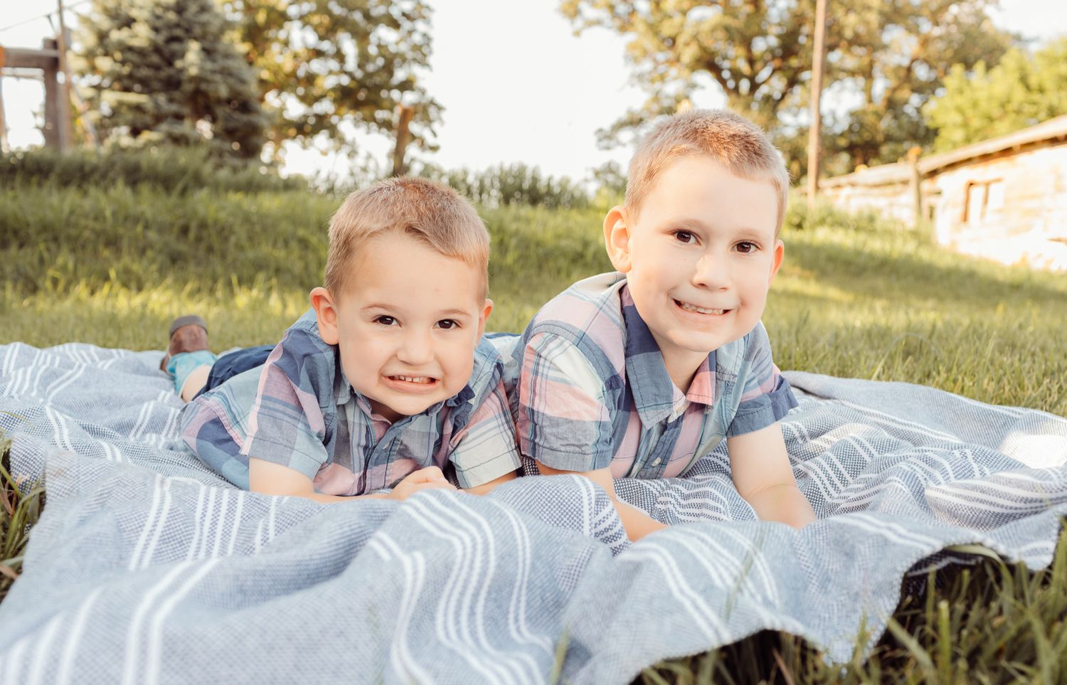 Two young brothers in matching plaid shirts smile while lying on a blanket in green grass outdoors.