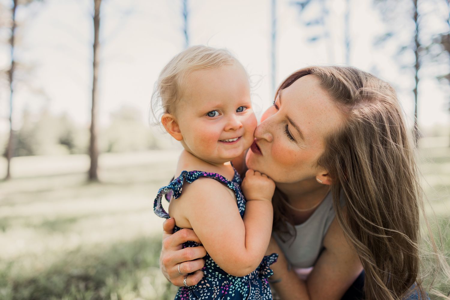 Young mother lovingly kisses her smiling toddler daughter outdoors in a sunny park setting.