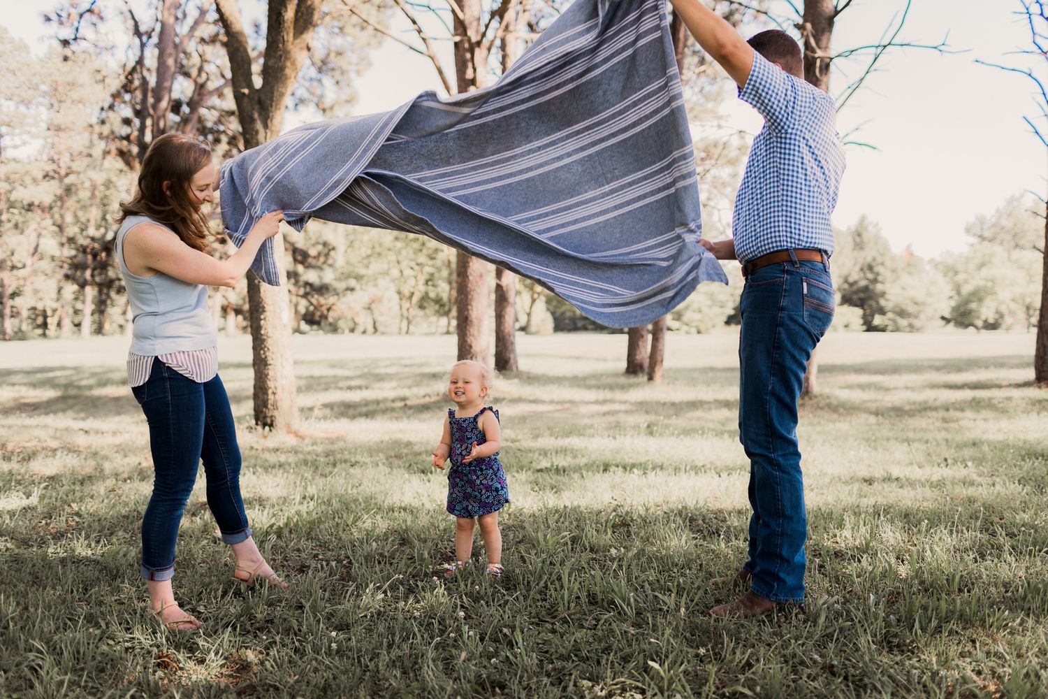 Parents playfully lift a striped blanket over their toddler daughter standing on green grass in a park.