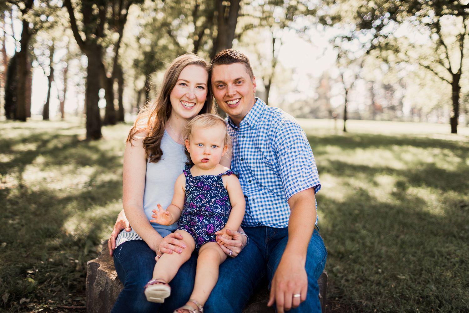 Smiling young family of three sits together outdoors on a log surrounded by lush green trees.