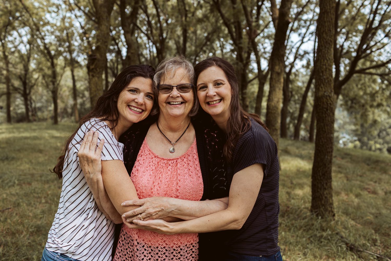 Three smiling women hug closely together outdoors in a wooded area with green trees behind them.