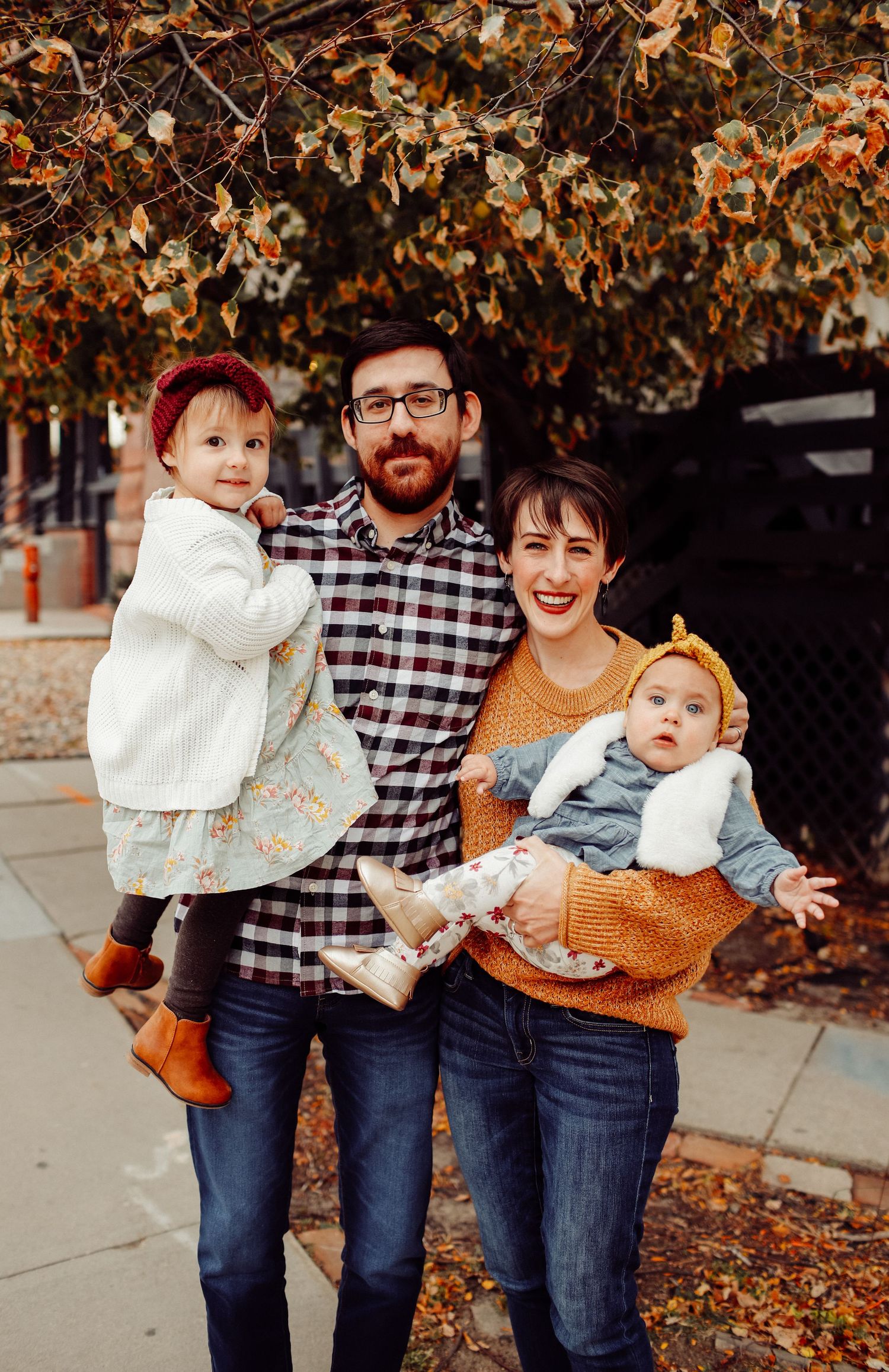 Happy family of four poses outdoors in fall, parents holding two young daughters in autumn clothing.