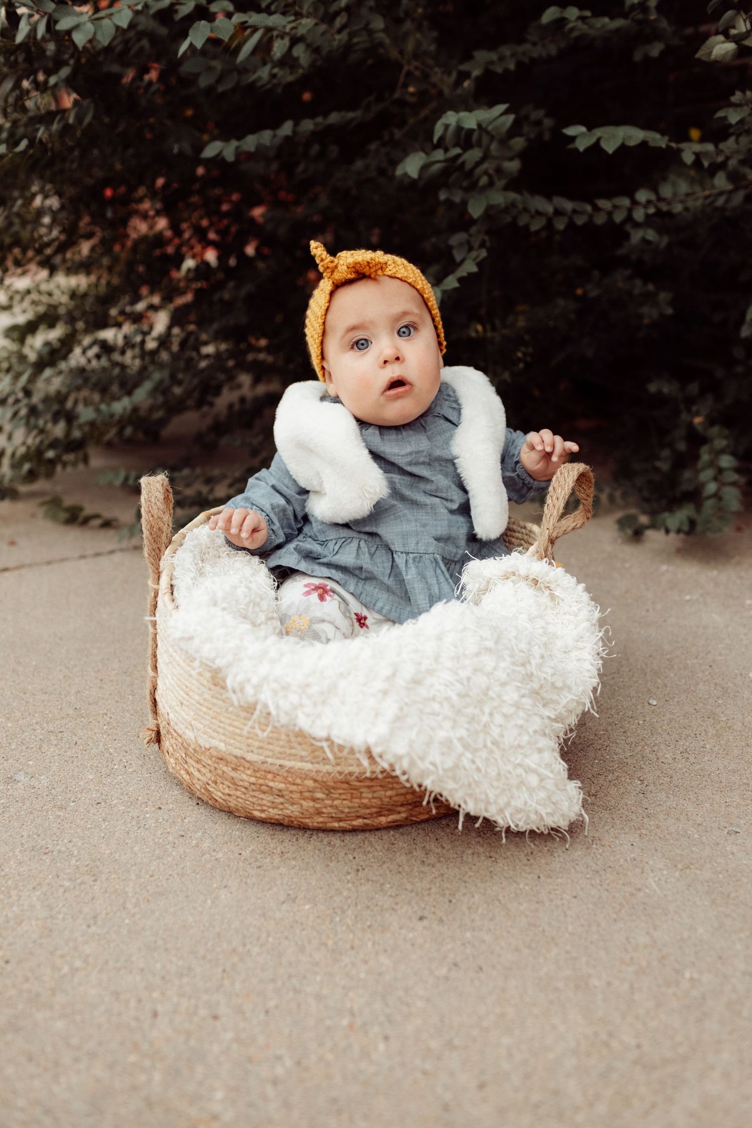 Adorable baby in yellow bonnet and gray dress sits in wicker basket with white fluffy blanket outdoors.
