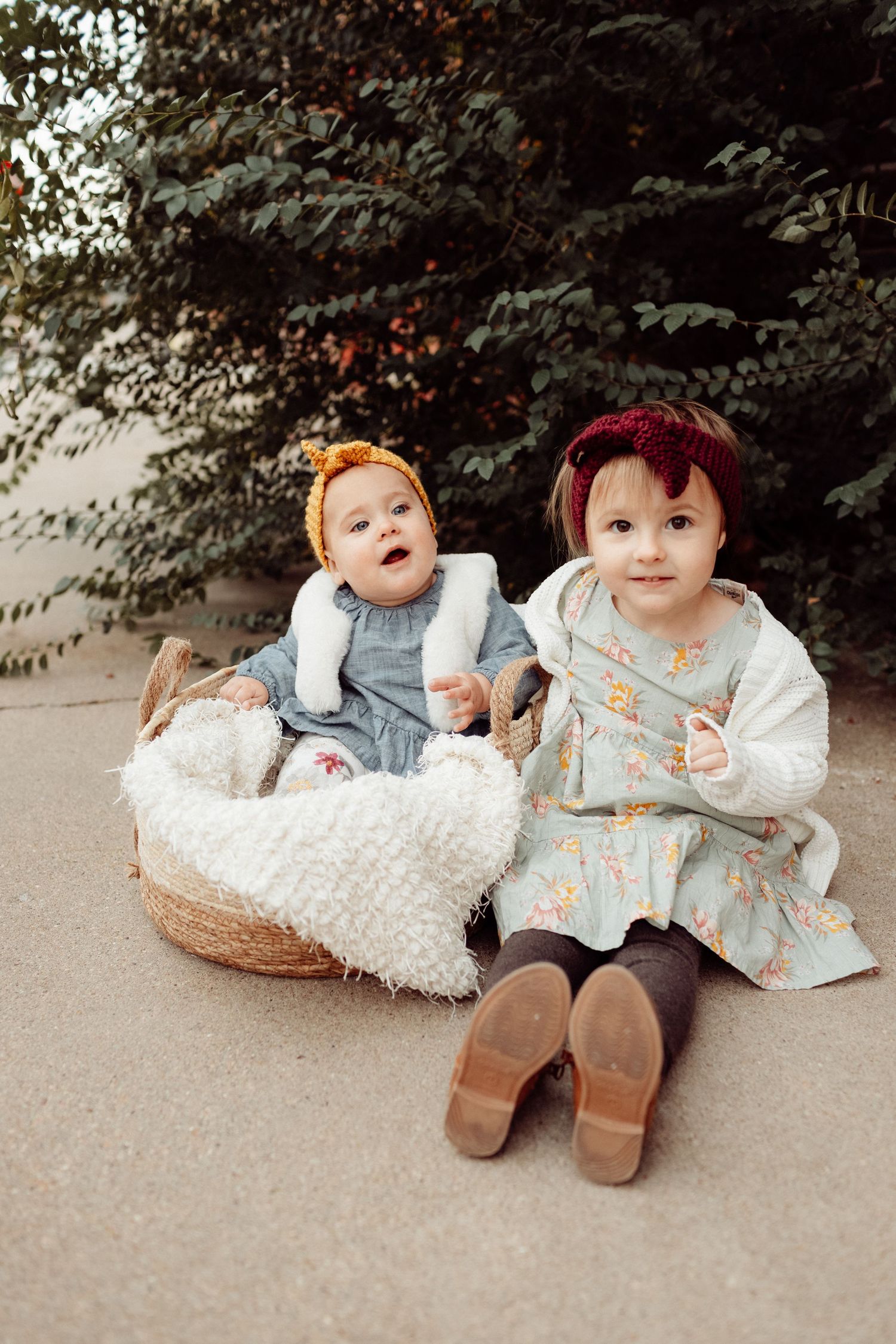 Two baby girls in colorful dresses sit outdoors by green trees, one in a wicker basket with white blanket.