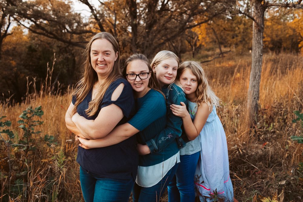 Smiling mother poses with her three daughters hugging her from behind in an autumn field setting.