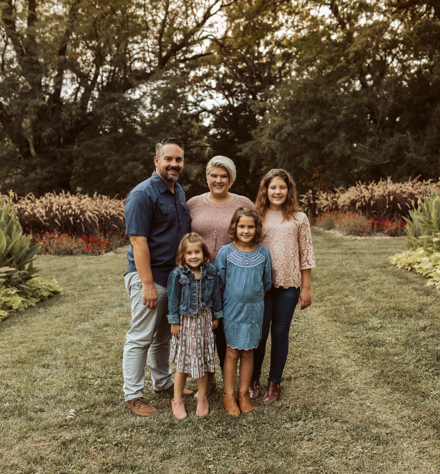Family of five smiles together on a lush green lawn surrounded by colorful autumn garden foliage.