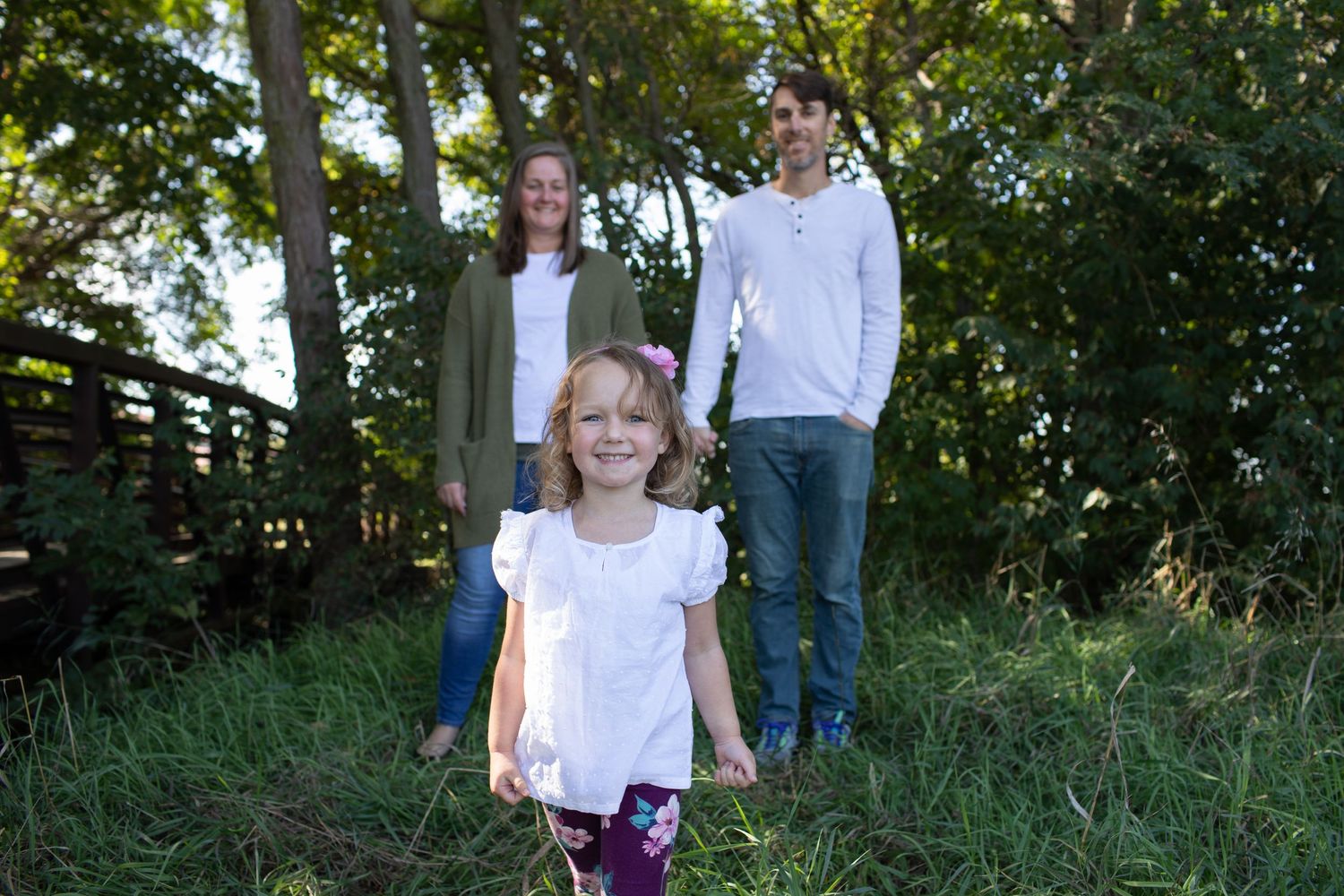 Family portrait with young girl in white shirt standing in lush green outdoor setting.