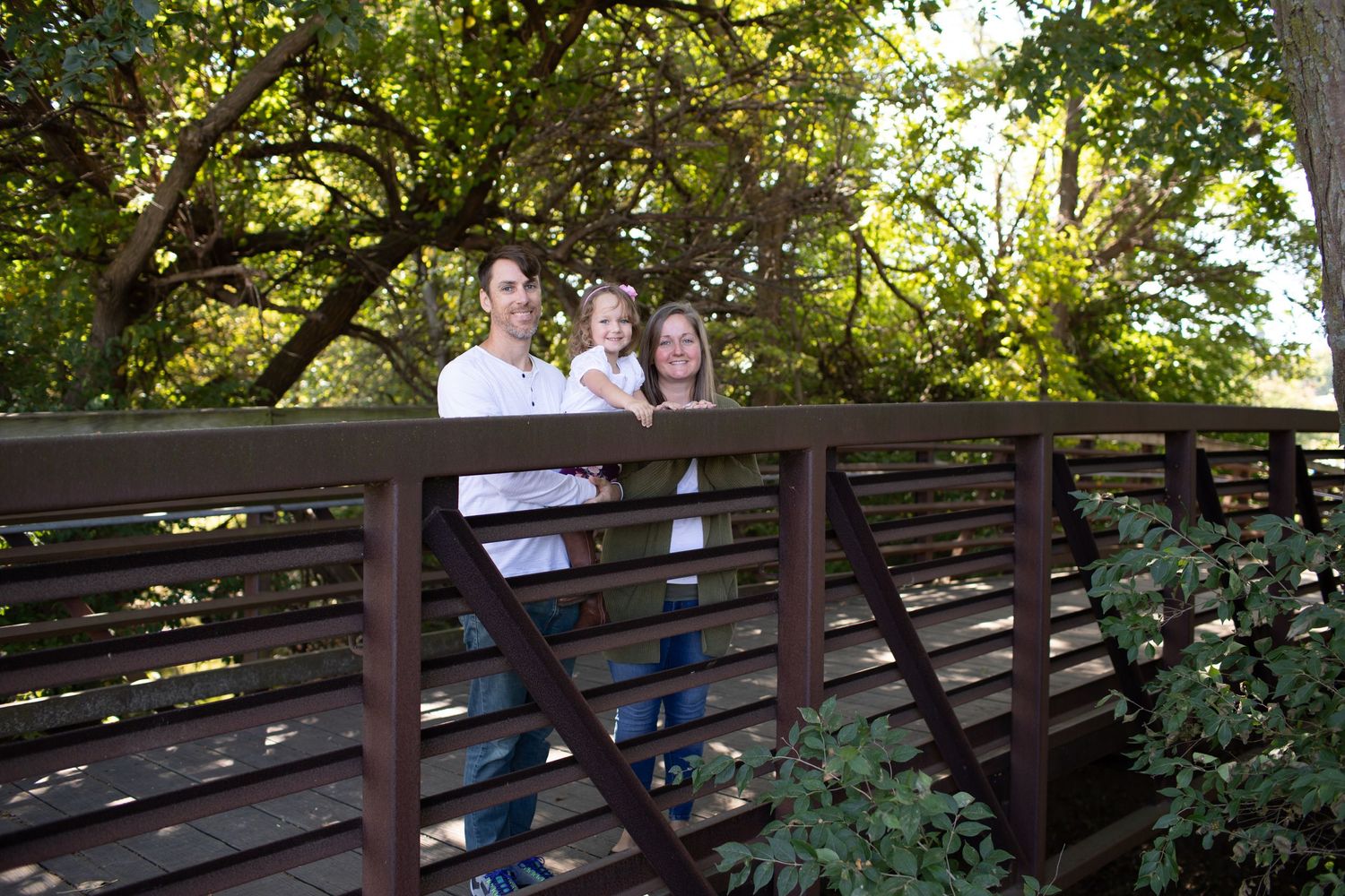 Happy family of three posing on wooden bridge surrounded by green trees.