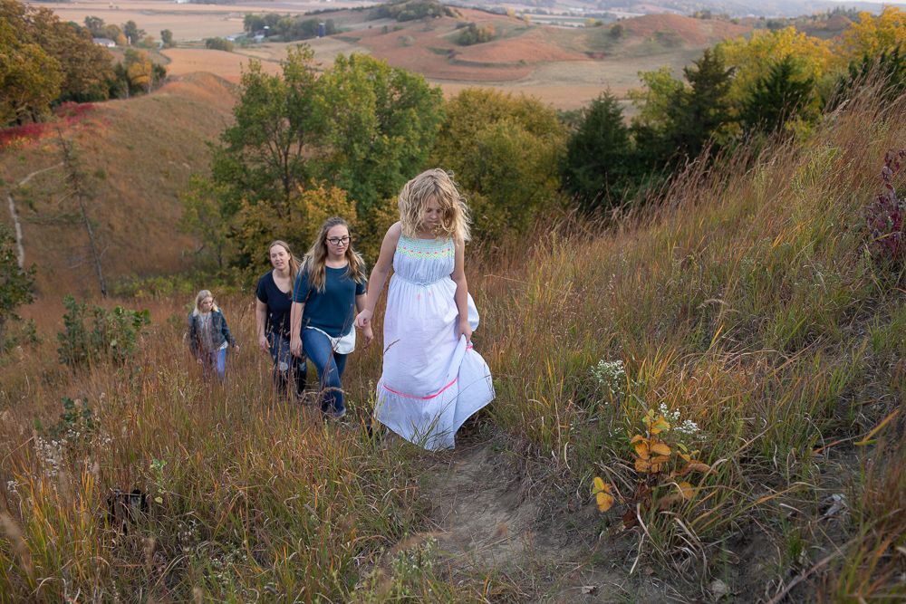 Women in white dress leads group hiking up scenic hill trail in autumn landscape.