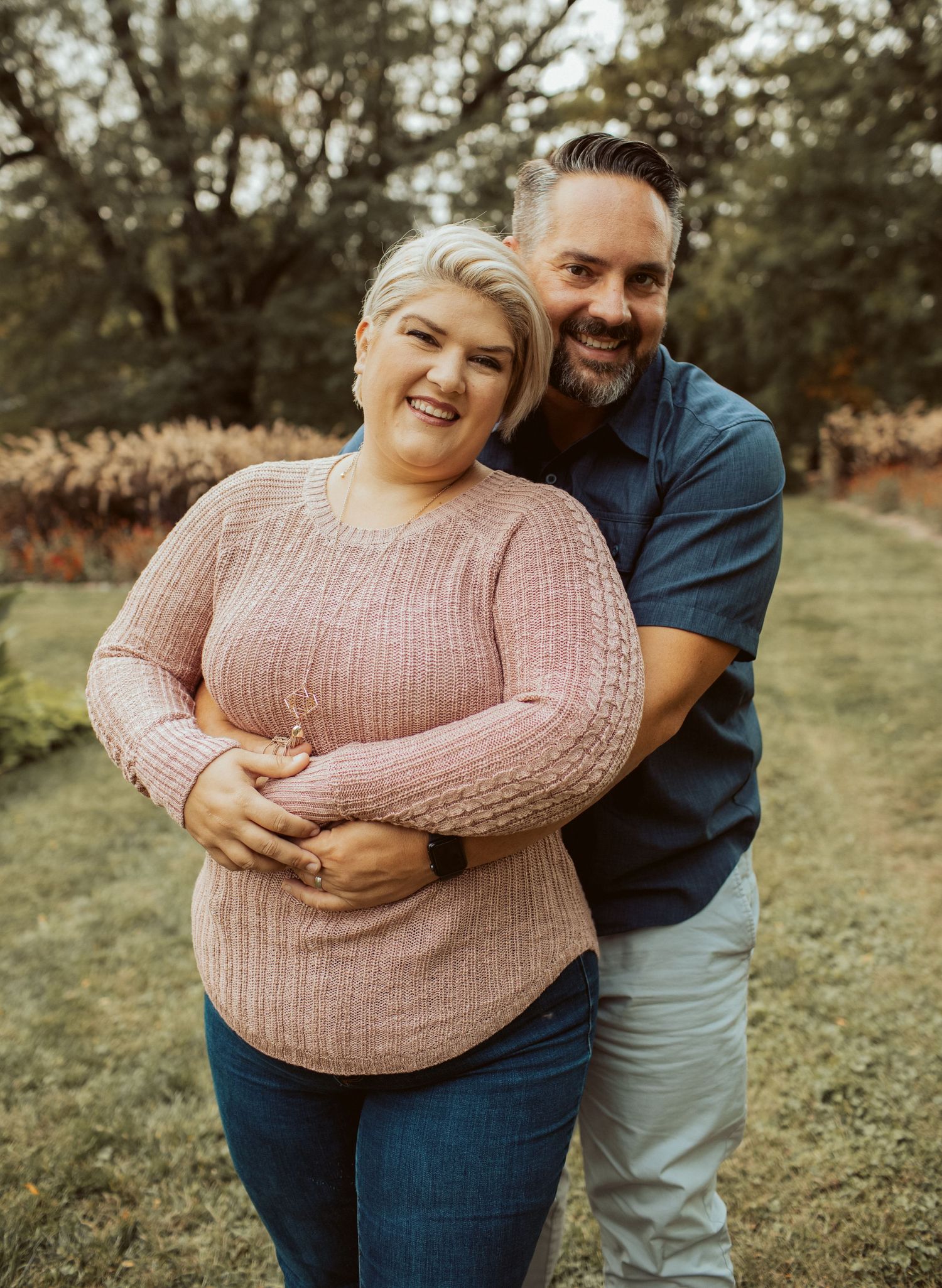 Smiling couple embraces outdoors in a garden setting, woman in pink sweater, man in blue shirt.