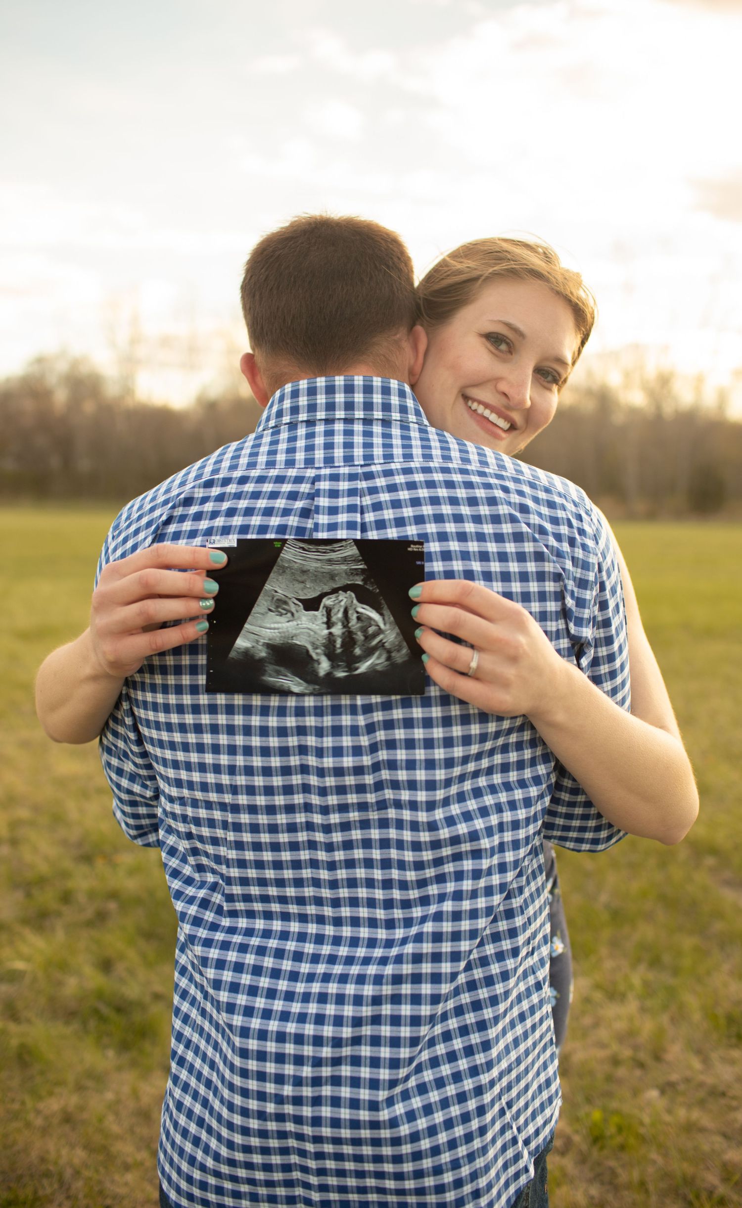 Smiling woman hugs man holding ultrasound photo in sunny field pregnancy announcement.