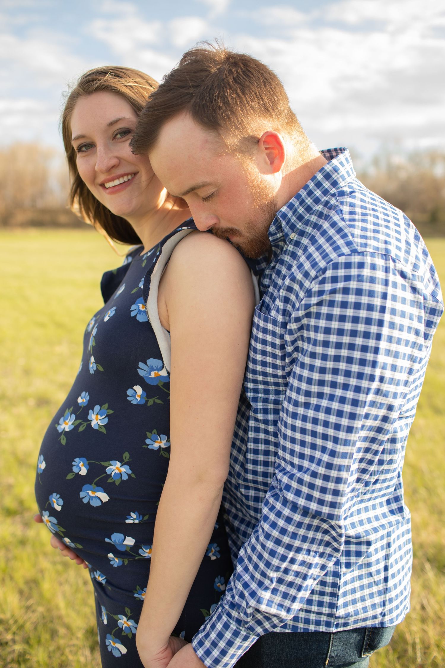 Pregnant couple embracing outdoors in sunny field, woman smiling in blue floral dress.