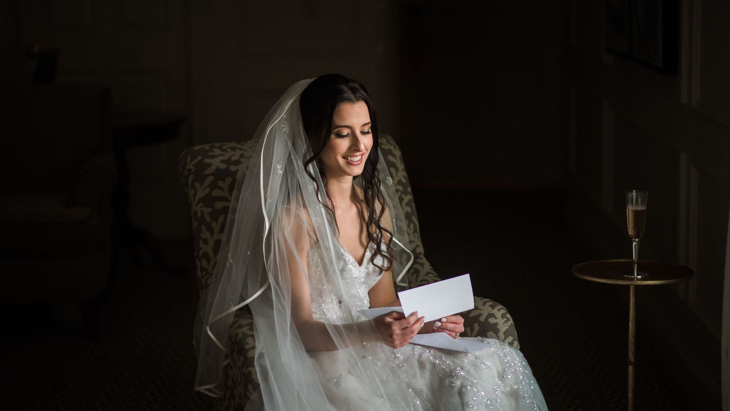 Radiant bride in lace wedding gown reads a heartfelt letter while seated in a chair.