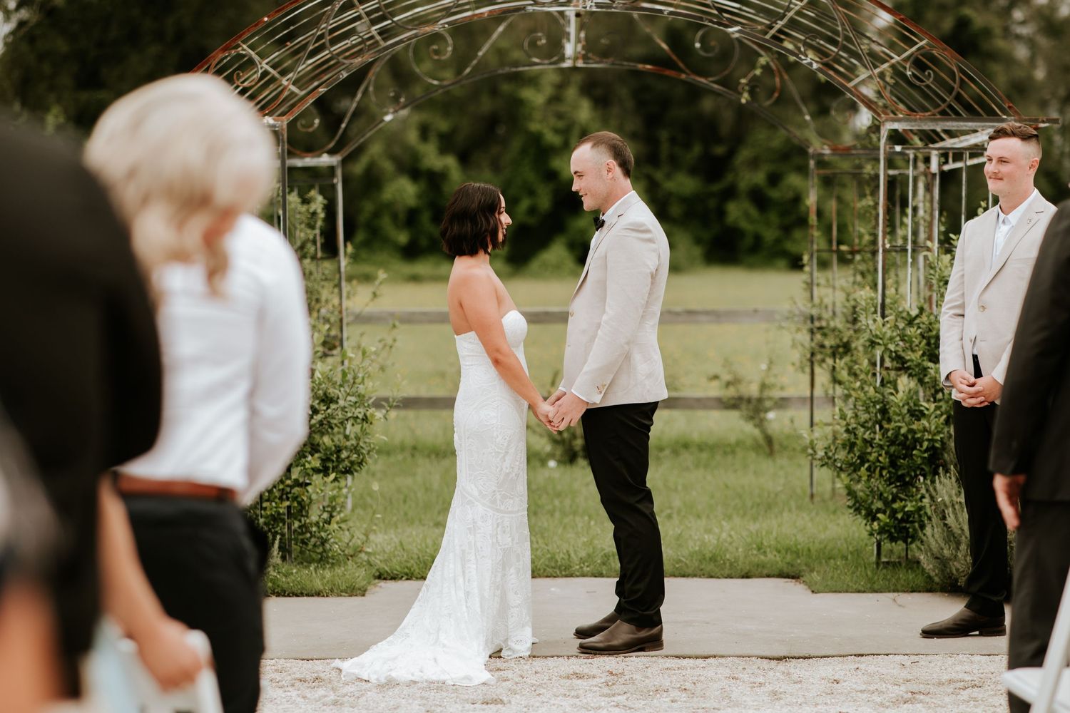 An outdoor wedding ceremony takes place under a rustic metal arch.