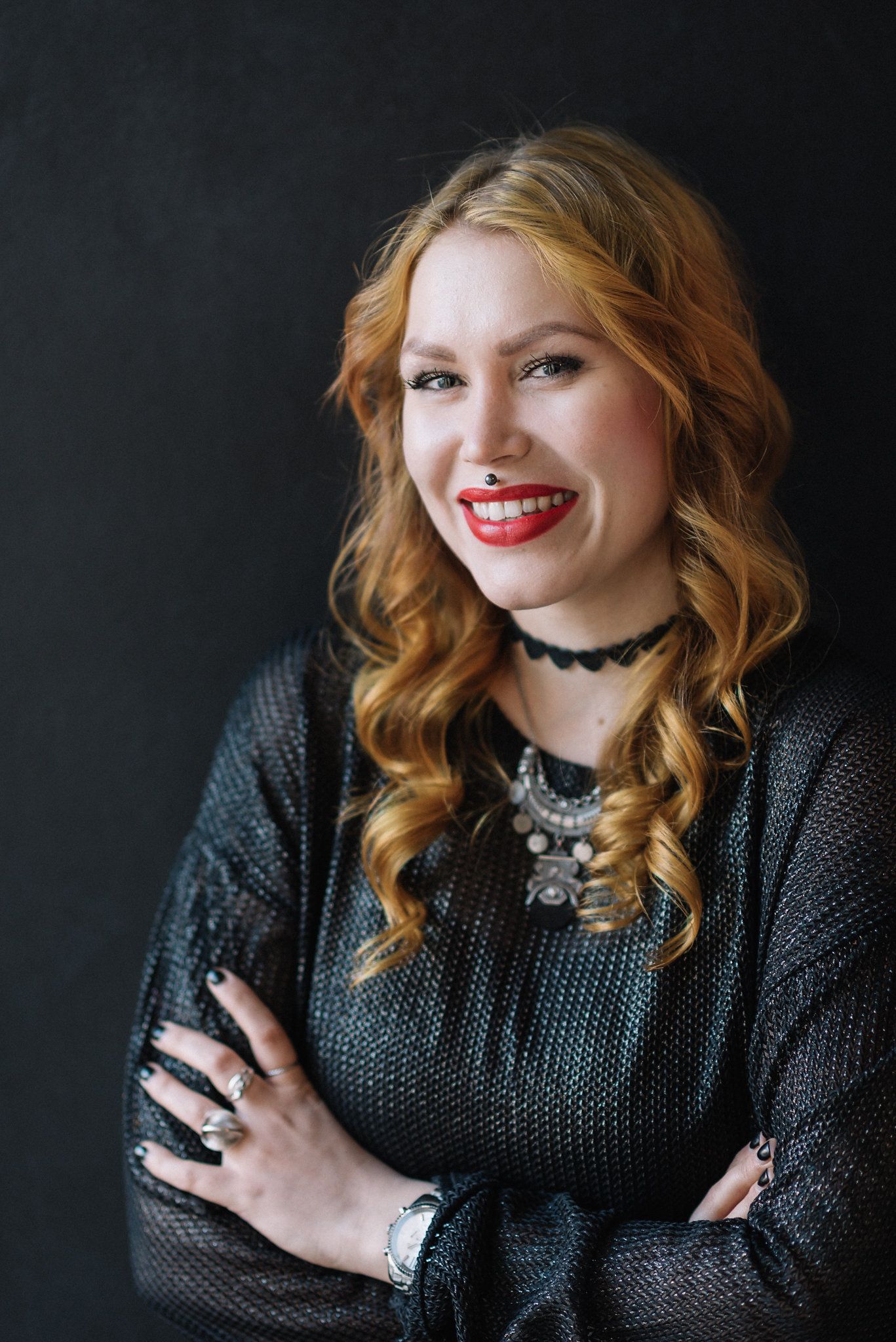 Professional studio portrait featuring wavy copper hair, dark sweater, statement necklace and choker against black background.
