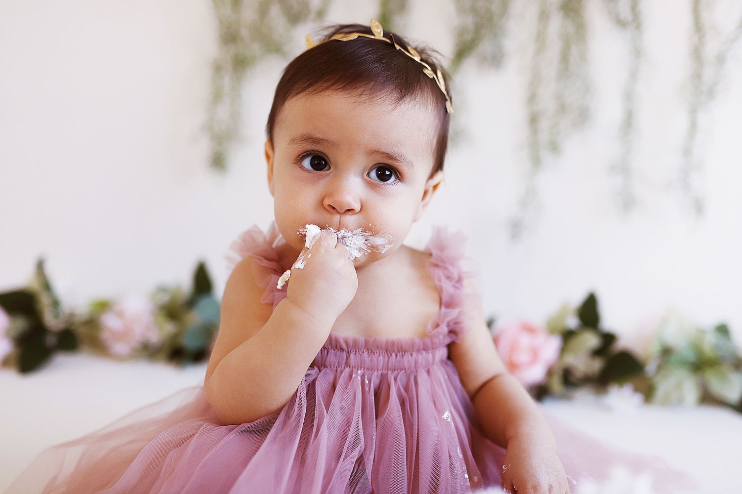 Baby in pink dress touching face with finger against floral backdrop.