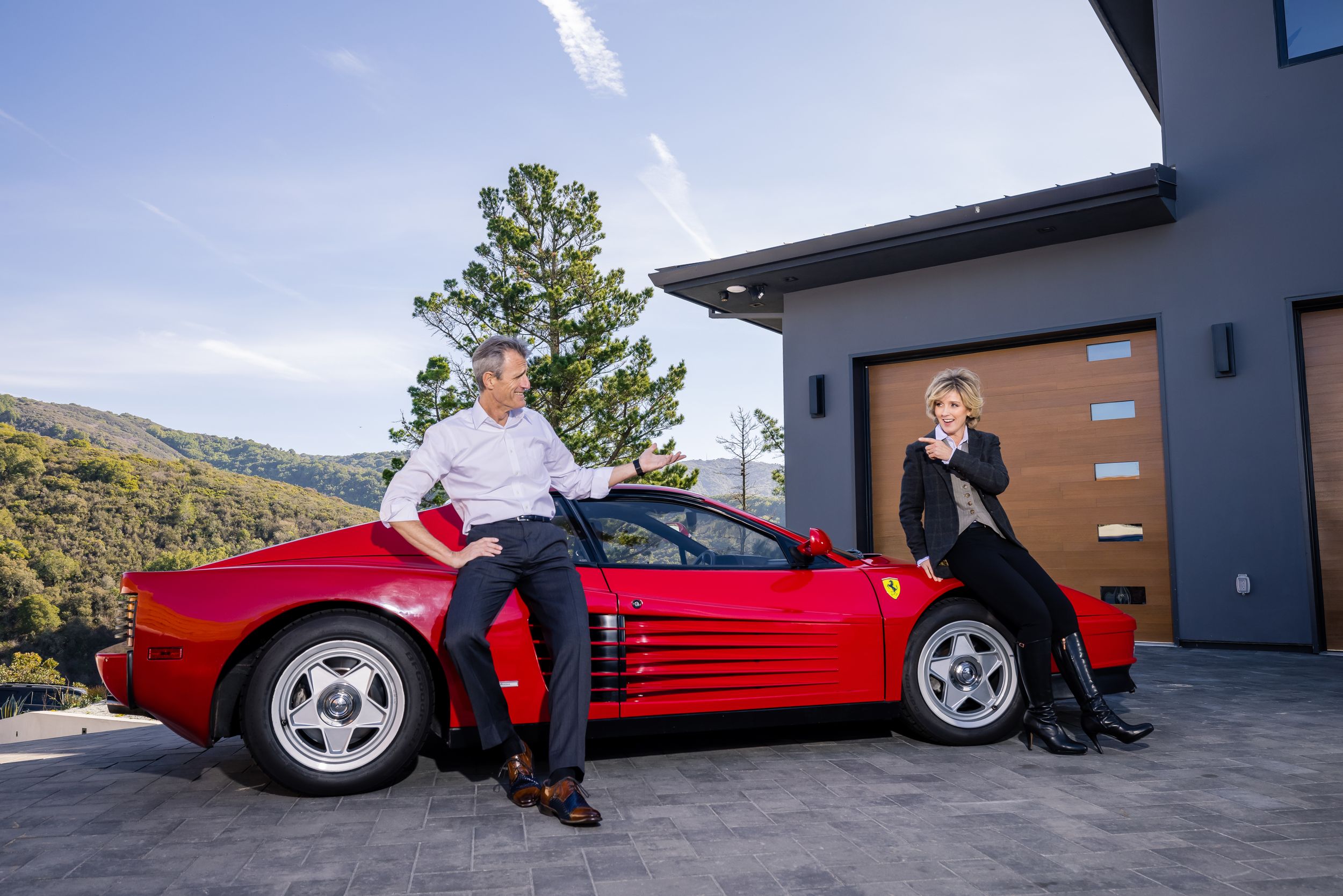 Man and a woman sitting on a red Ferrari smiling and pointing at each other