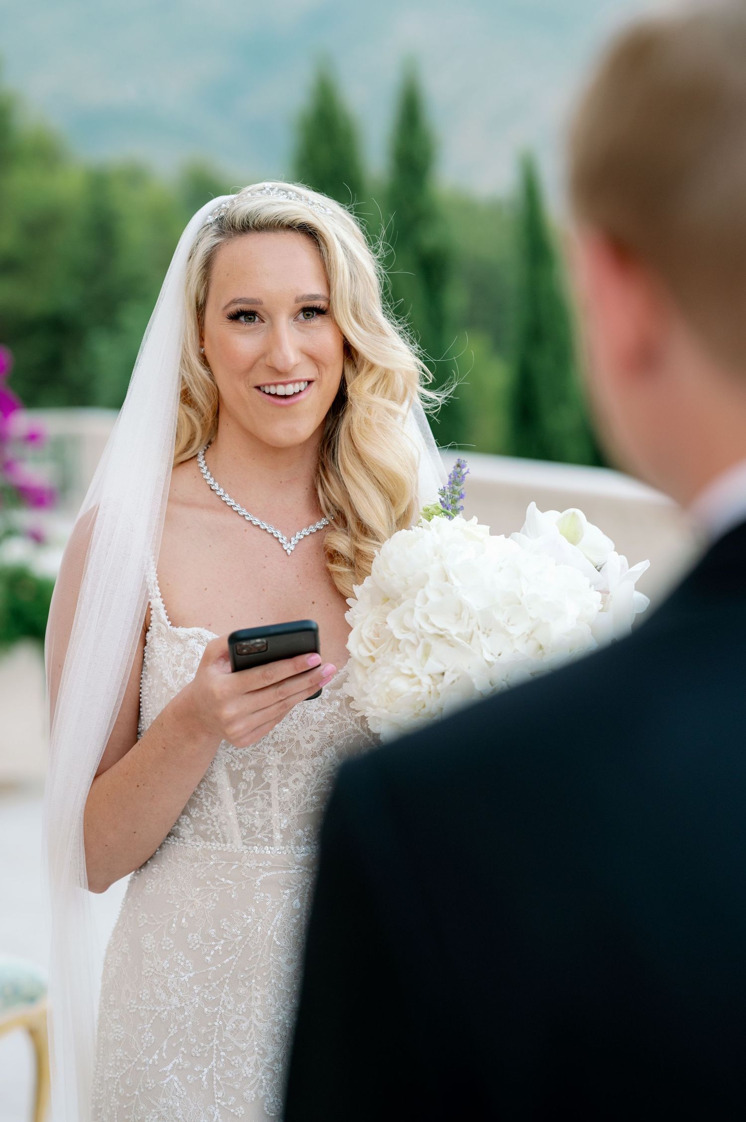 Bride in lace gown holds phone while carrying white bouquet during outdoor ceremony.
