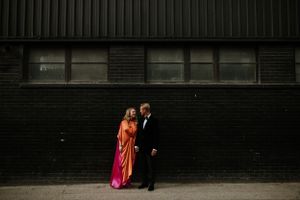A couple in formal attire stands against a dark industrial building wall, with the woman wearing a vibrant pink gown in London.