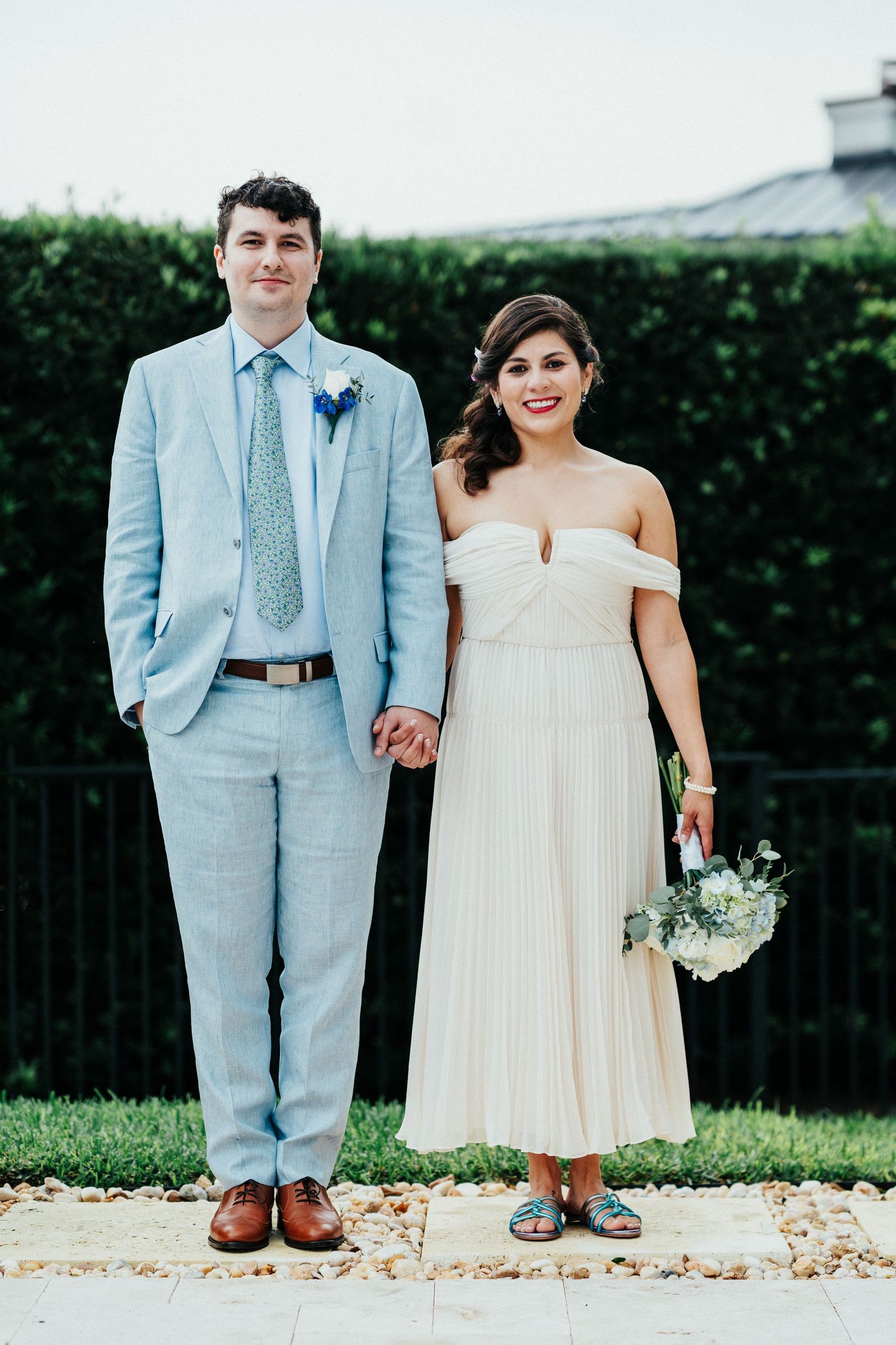 Newlyweds in light blue suit and off-shoulder white dress pose together in garden setting.