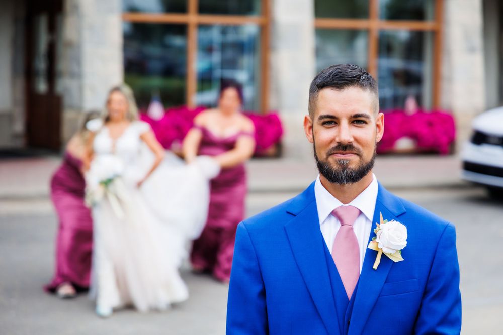 A wedding party poses outside with bridesmaids in magenta dresses and the groom in a bright blue suit.