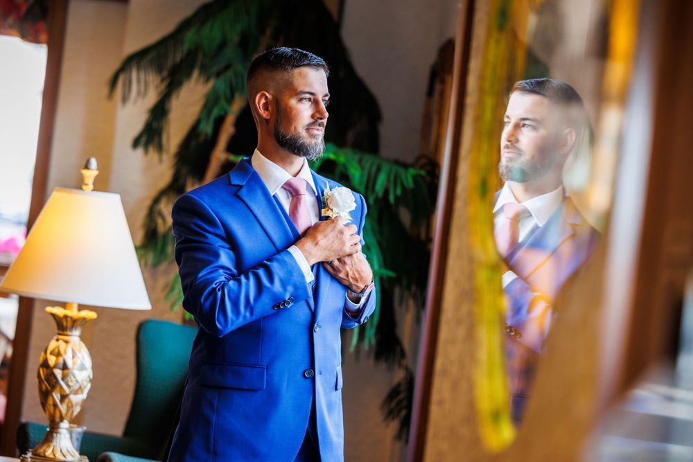 A formal event preparation scene showing a groom in a bright blue suit adjusting their tie in front of a mirror.