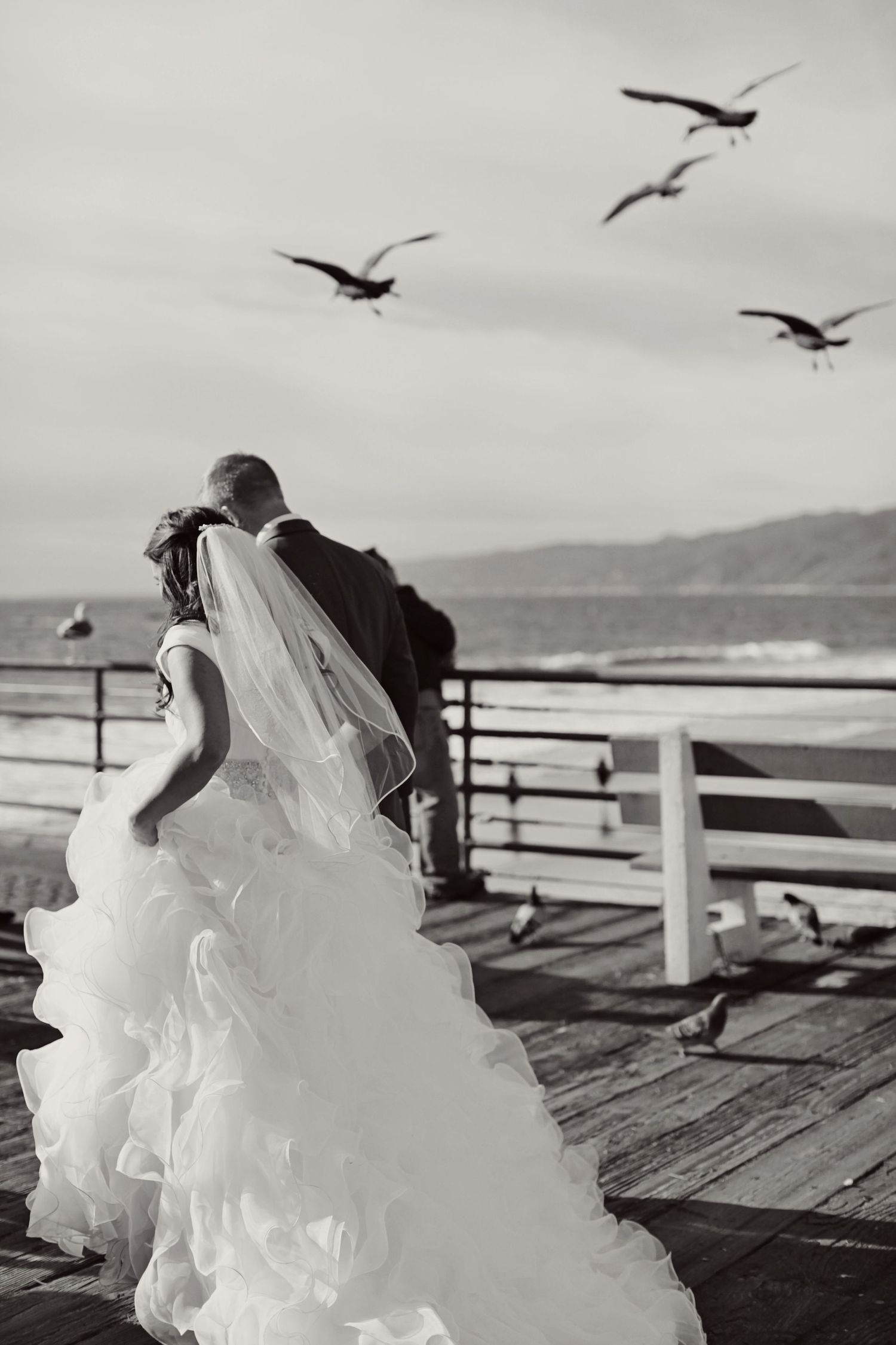 A bride and groom watch seagulls fly overhead on a wooden pier by the ocean.