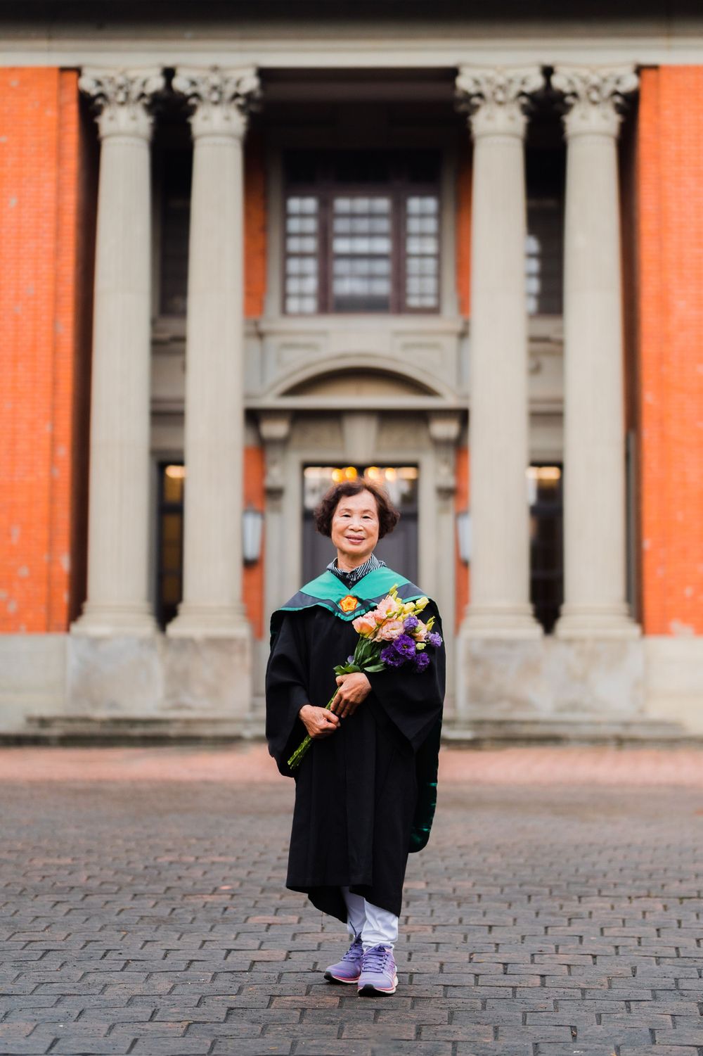 Graduate in black regalia and colorful bouquet poses with bright smile in front of university columns.