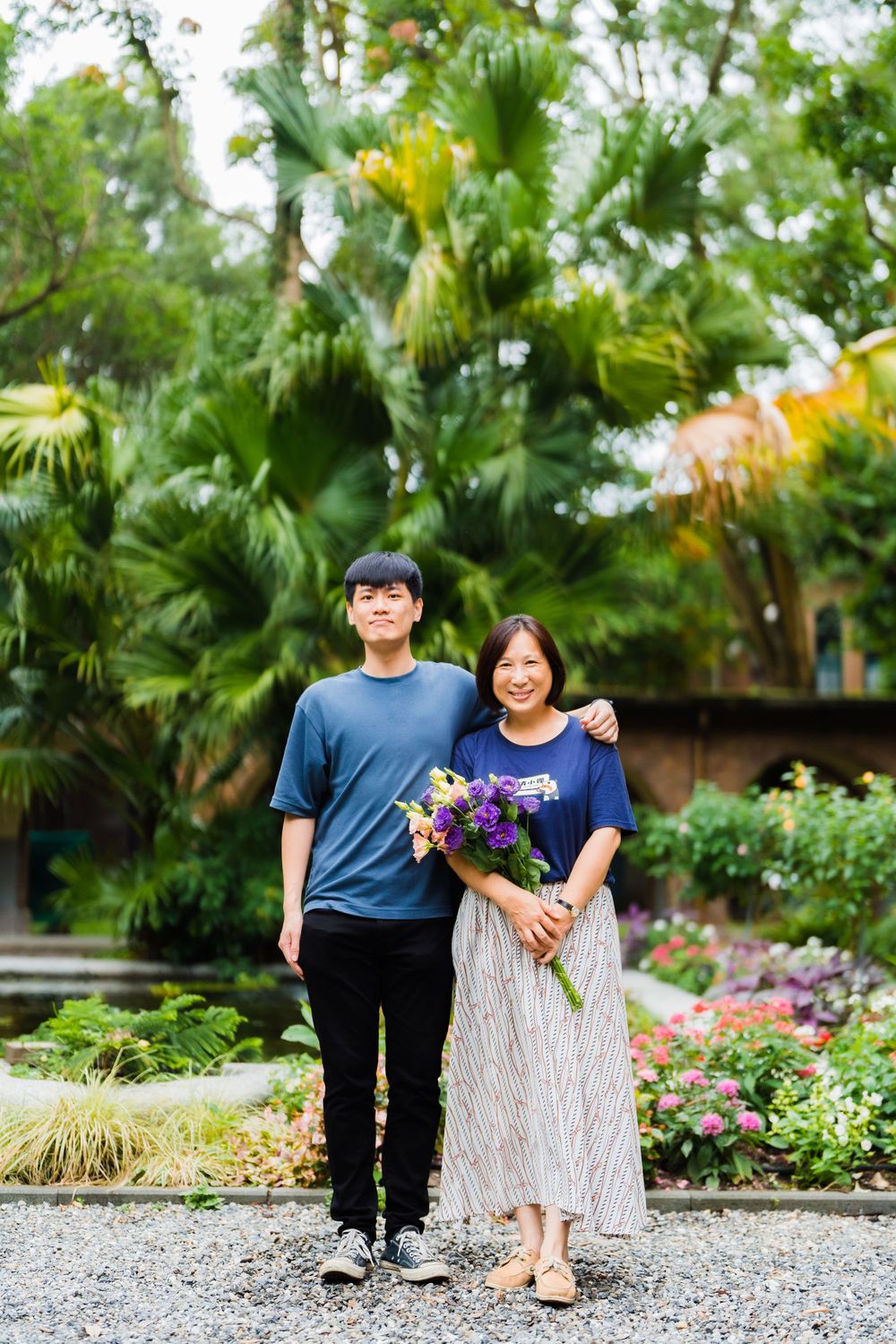 A couple poses together with purple flowers in a tropical garden setting.