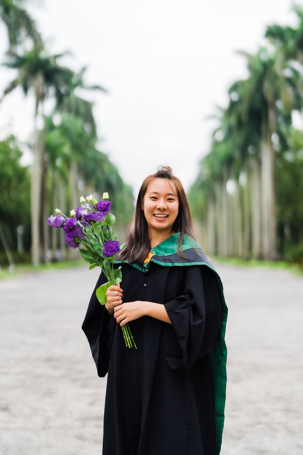 Graduate in black academic regalia holds purple flowers while posing on palm-lined walkway.