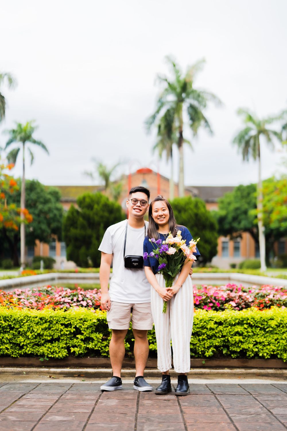 Couple posing with flowers in front of palm trees and a red-roofed building.