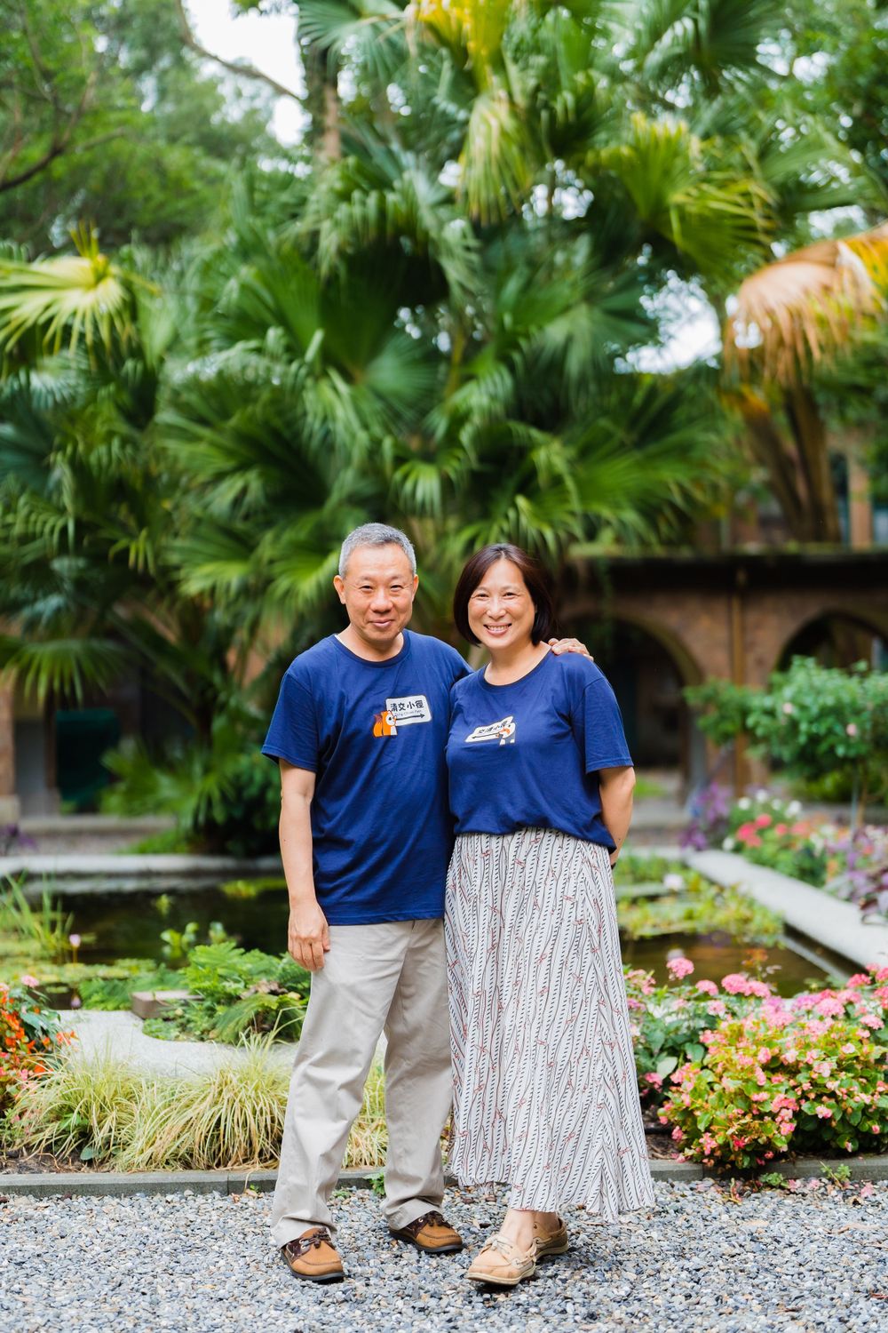 A couple in matching blue shirts poses together in a garden with palm trees.