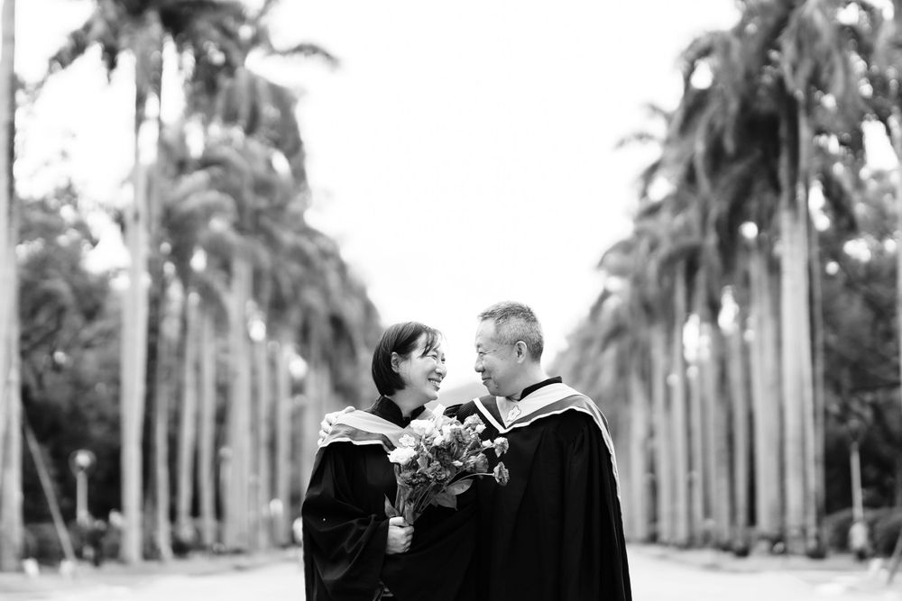 Black and white portrait of two people in graduation robes among palm trees.