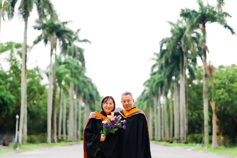 Graduation photo of graduates standing on a palm-lined path wearing black and orange academic regalia.