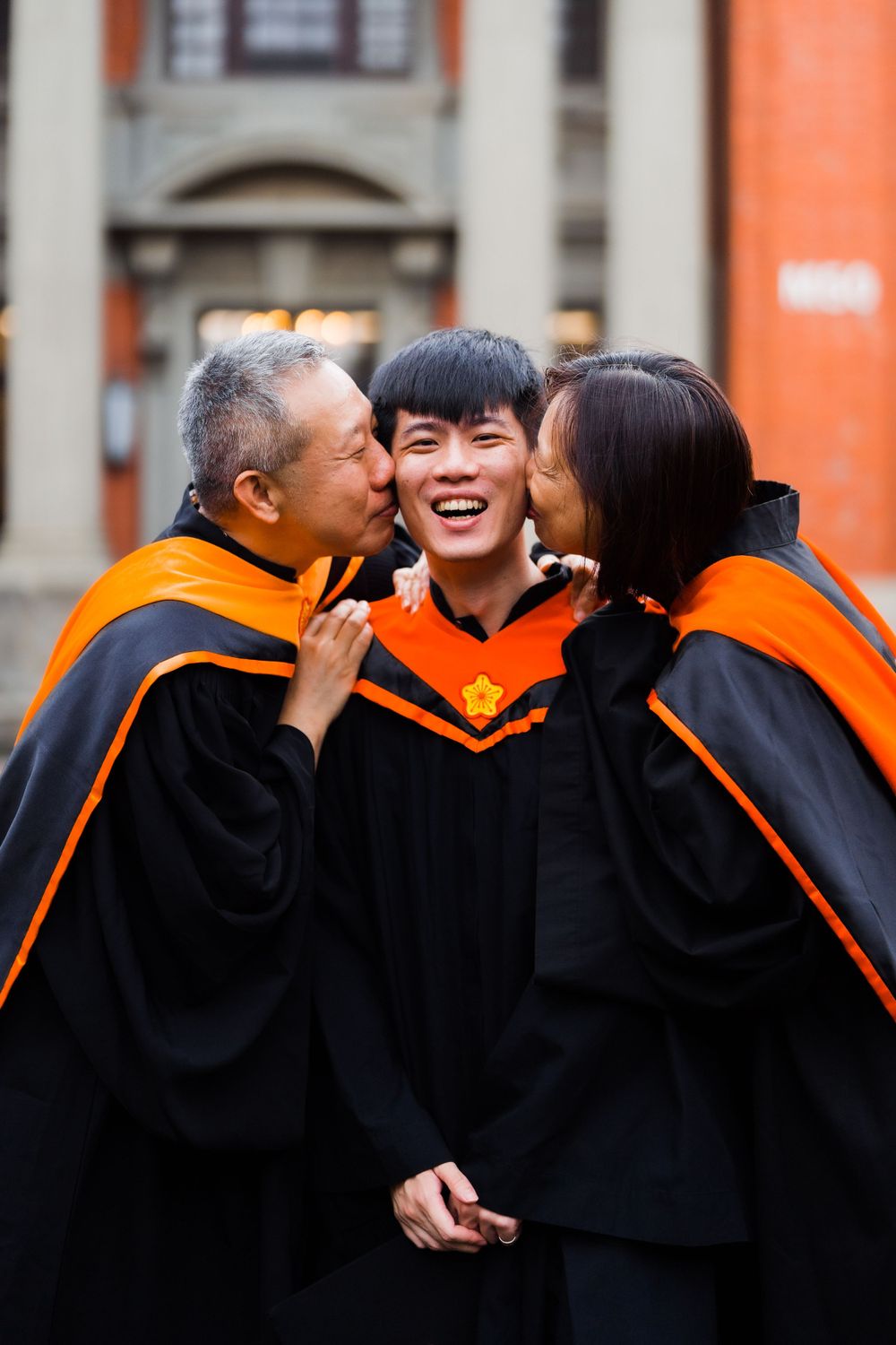 Three people in graduation robes share an intimate celebratory moment outside a university building.