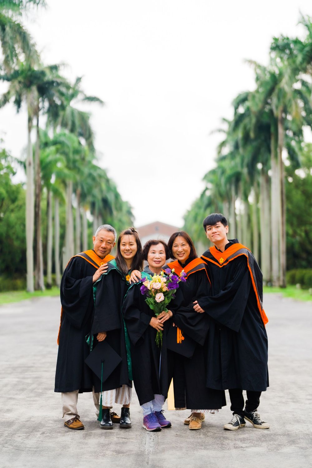 Graduation group photo with professors and graduate holding flowers on palm-lined path.