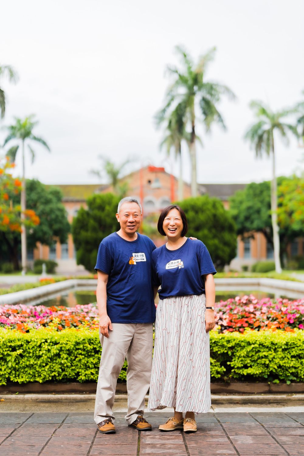Two people in matching blue shirts standing in front of palm trees and flowerbeds.