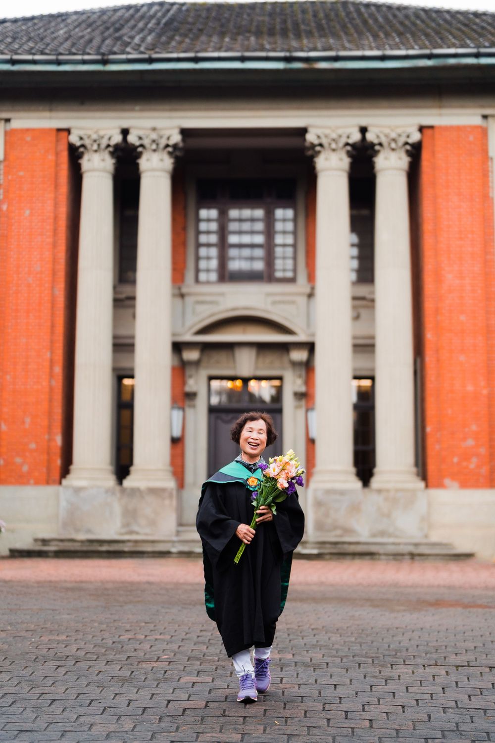 Graduate holding flowers walks confidently in front of ornate university building with columns.