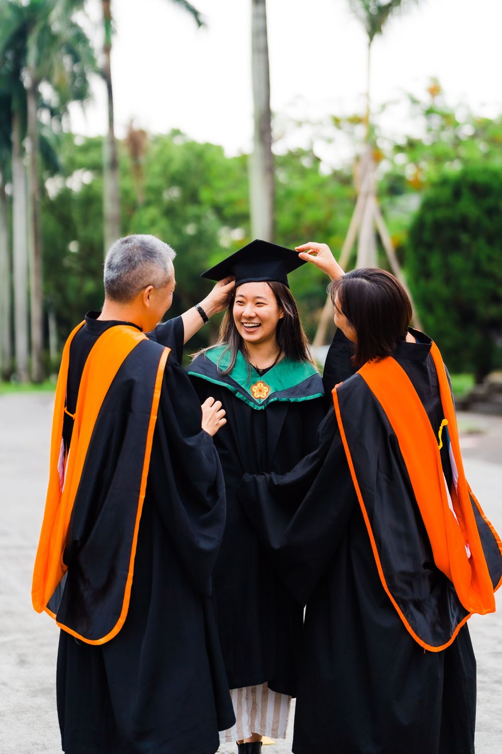 Graduate in traditional black and green regalia celebrating with faculty in orange-trimmed robes on campus.